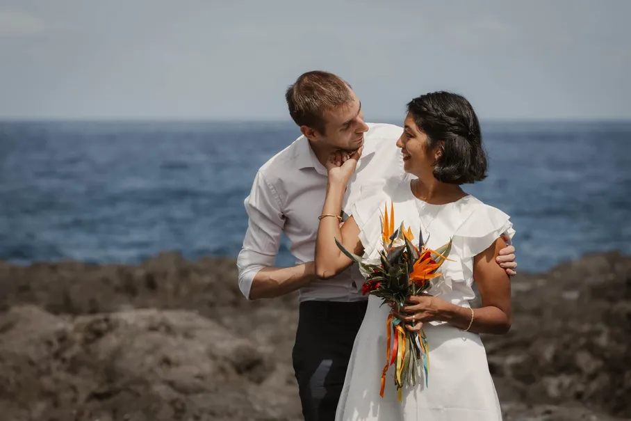 Un couple enlacé sur les rochers noirs face à l’océan, elle sourit, un bouquet coloré à la main