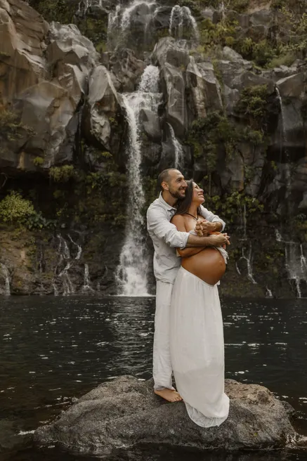 Un couple enlacé sur un rocher au pied d’une cascade, la future maman en jupe blanche