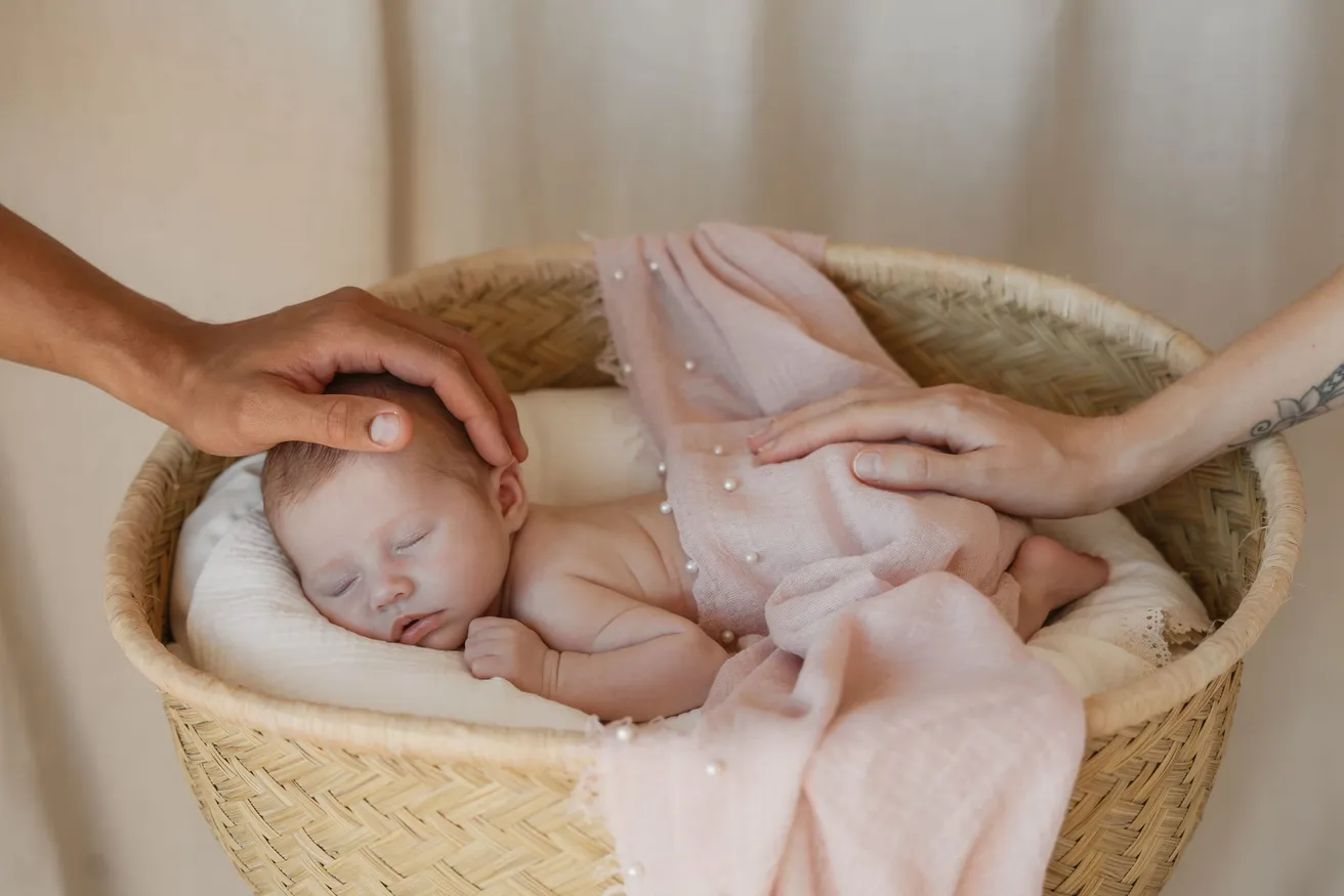Deux mains d’adultes posées sur le nouveau-né endormi dans un panier tressé, sous un voile rose