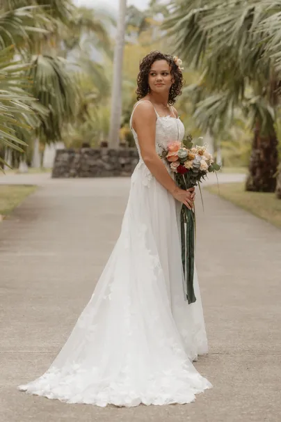 La mariée seule dans une allée de palmiers, bouquet pêche à la main, longue robe de dentelle