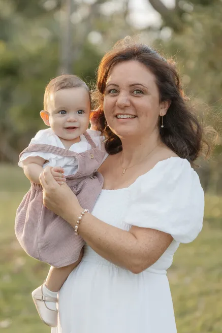 Une mère en robe blanche sourit en portant sa petite fille dans ses bras, devant la verdure