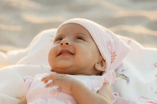 Bébé qui sourit, coiffée d’un bonnet fleuri, sur une couverture claire