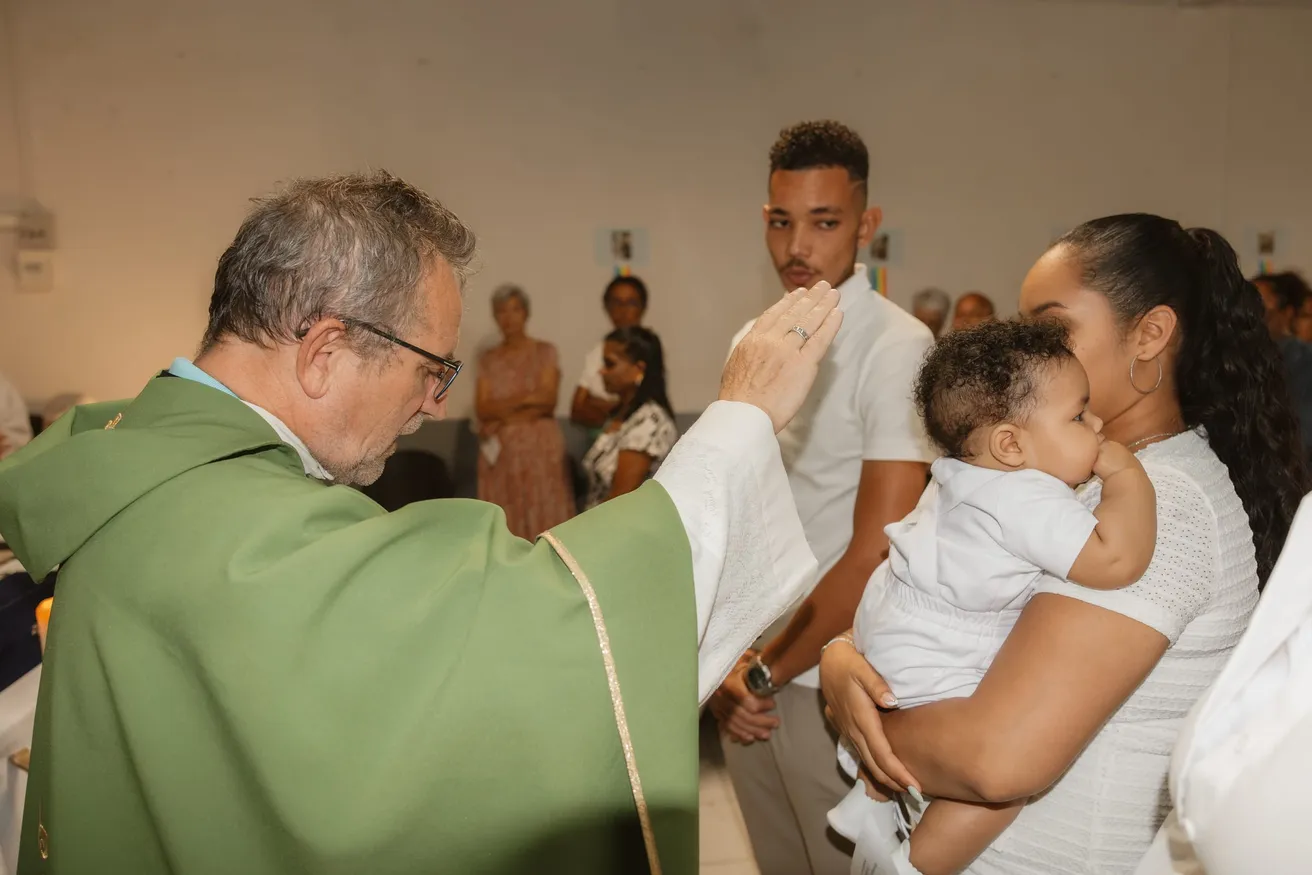 Le prêtre en chasuble verte bénit l’enfant porté par une jeune femme, devant l’assemblée