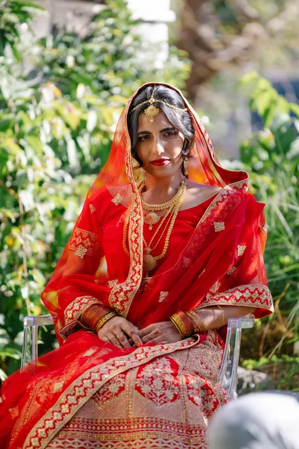 La mariée en sari rouge brodé d’or, parée de bijoux traditionnels, assise dans un jardin
