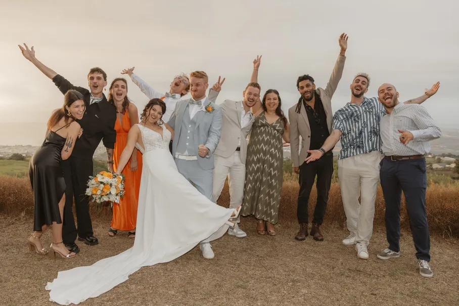 Les mariés et leurs amis lèvent les bras en l’air dans un champ, sous un ciel gris