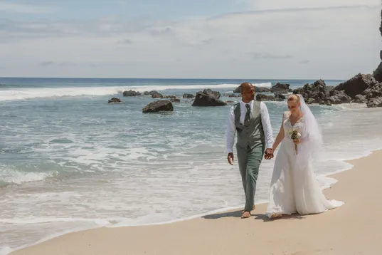 Les mariés marchent main dans la main sur le sable, au bord de l’eau et des rochers