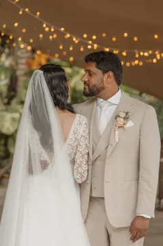 Le marié regarde la mariée de dos sous les guirlandes lumineuses, voile sur la dentelle