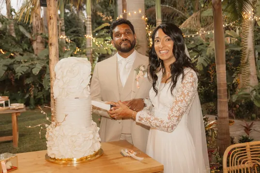Les mariés coupent ensemble un gâteau blanc à volants dans un jardin tropical