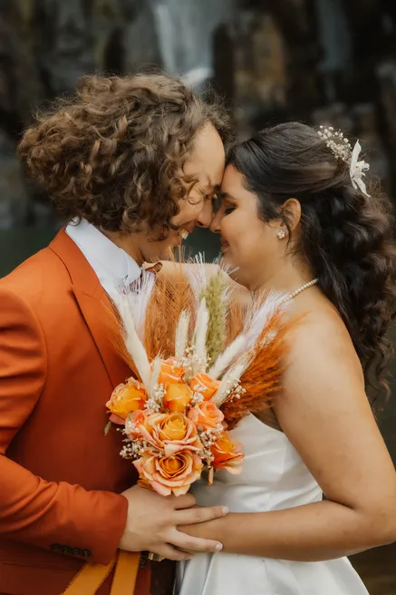 Les mariés front contre front, veste rouille et bouquet de fleurs séchées orange entre eux