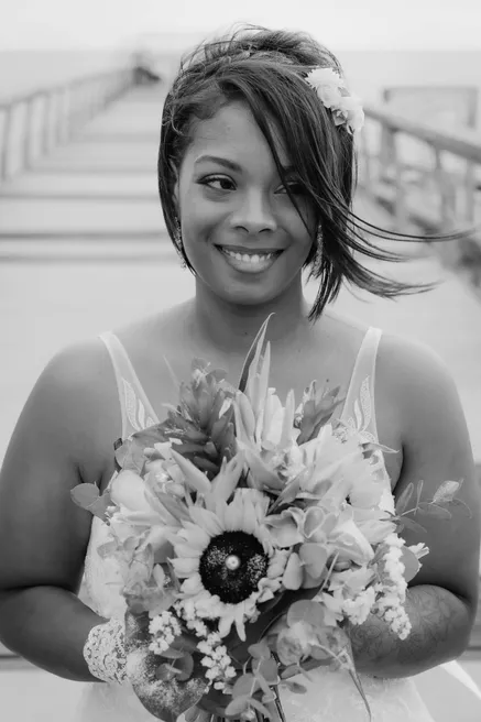 En noir et blanc, la mariée sourit, cheveux au vent, un bouquet de tournesol contre elle