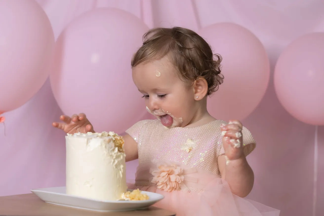 Une petite fille en robe rose tire la langue en attrapant la crème de son gâteau blanc