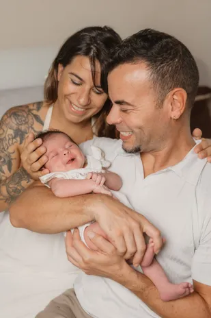 Un couple rit en tenant son nouveau-né, le père enlaçant la mère aux bras tatoués