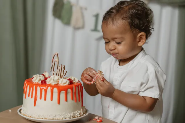 Un petit garçon en chemise blanche prend un morceau de son gâteau à la crème orange, sous la guirlande « 1 »