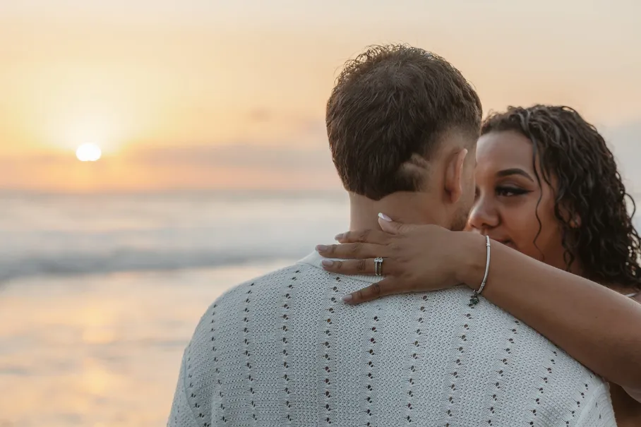 Une femme enlace le cou de son compagnon vu de dos, face au soleil qui se couche sur la mer