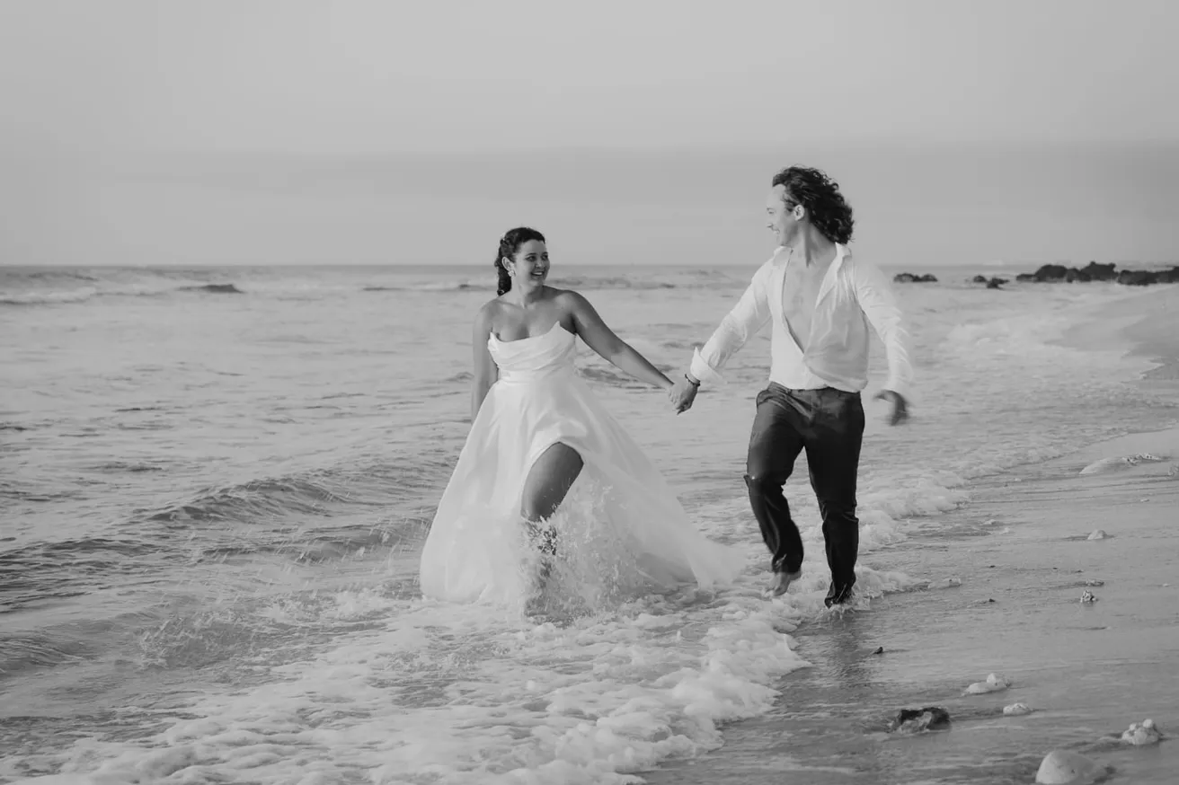 En noir et blanc, les mariés courent dans les vagues en se tenant la main