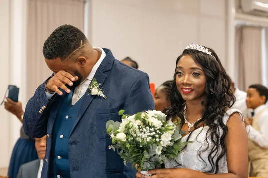 Le marié essuie une larme à côté de la mariée souriante qui tient un bouquet blanc