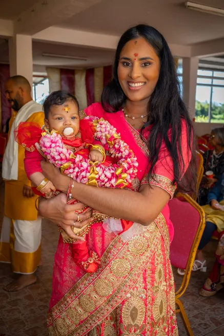 Femme en sari rose et doré portant le bébé en tenue rouge et guirlande de fleurs