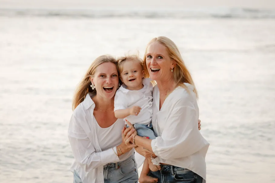 Deux mamans éclatent de rire en tenant leur petite fille, au bord de la mer