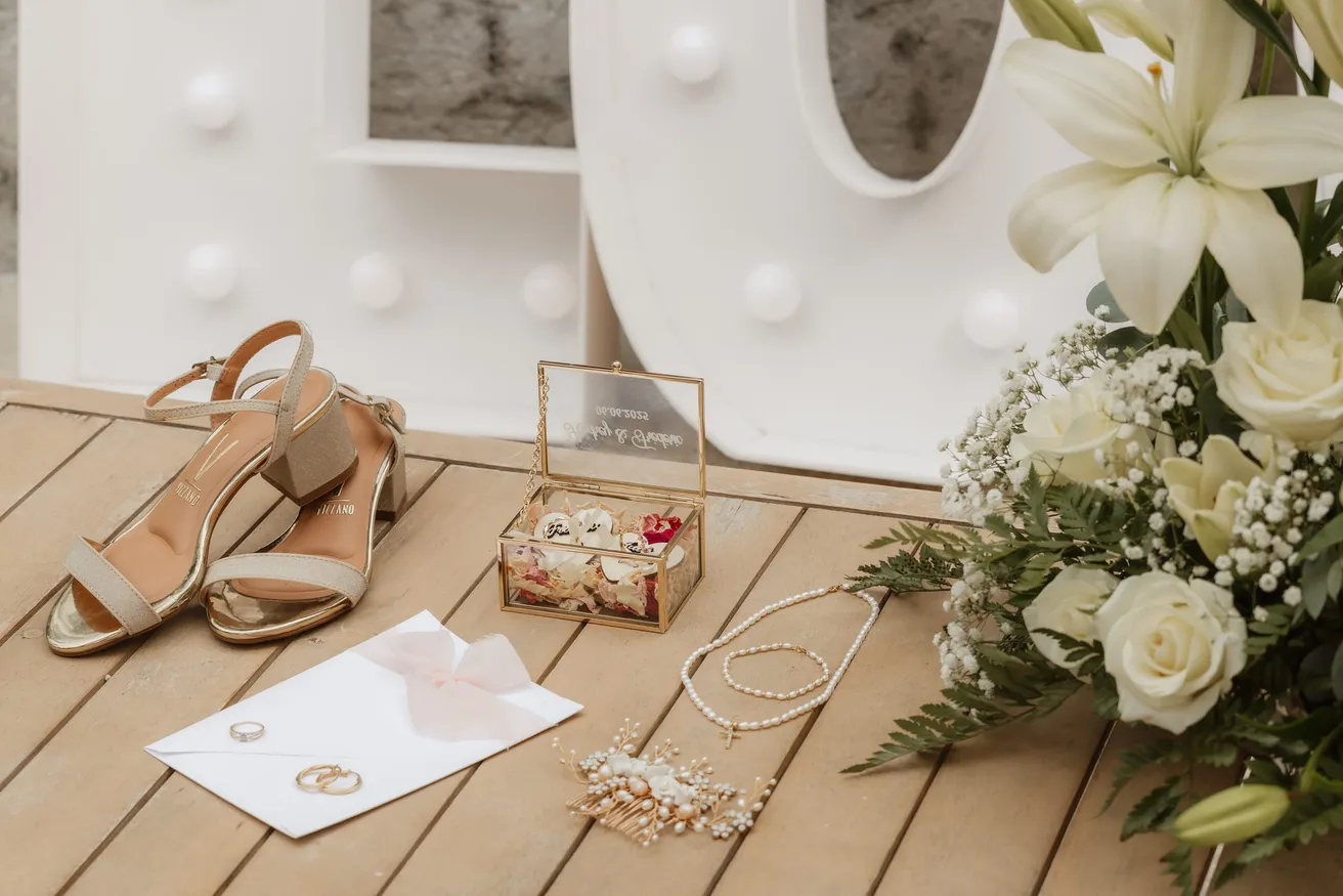 Sandales dorées, coffret à alliances, bijoux et enveloppe posés sur un plancher, près de lys blancs