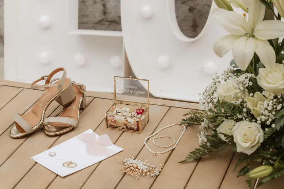 Sandales dorées, coffret à alliances, bijoux et enveloppe posés sur un plancher, près de lys blancs
