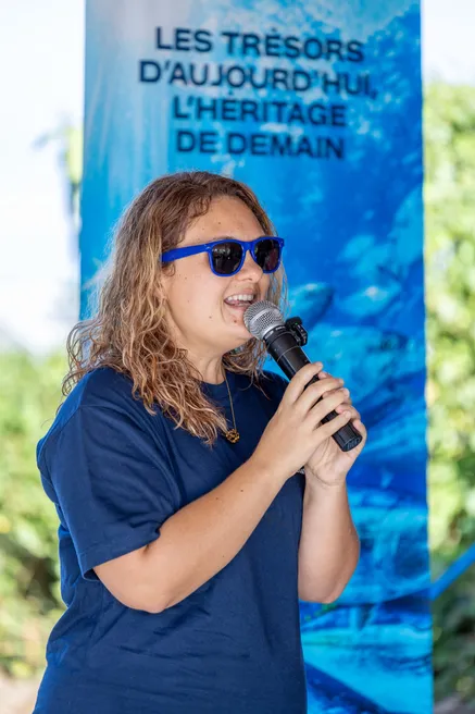 Une femme en tee-shirt bleu et lunettes de soleil parle au micro devant une banderole