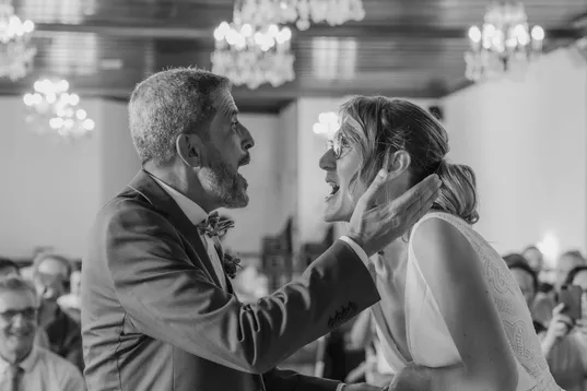En noir et blanc, un homme aux cheveux gris prend le visage de la mariée dans ses mains, sous des lustres