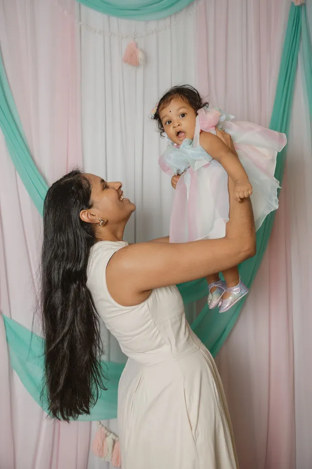 Une maman en robe blanche soulève sa fille en robe pastel devant le décor menthe et rose