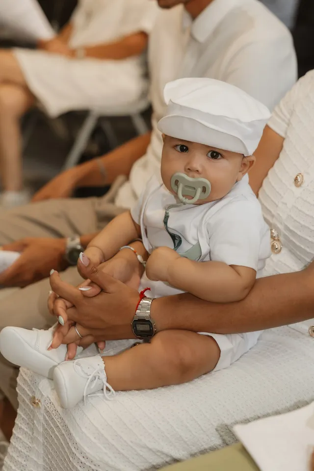Un bébé en ensemble blanc et casquette, assis sur les genoux d’un proche, une tétine accrochée à sa tenue