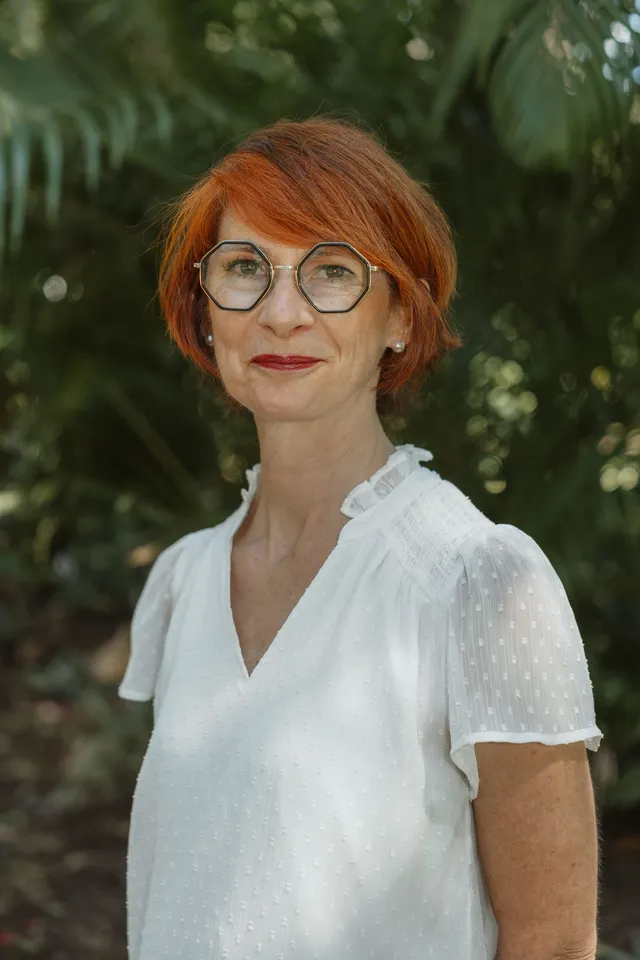 Une femme rousse à lunettes, en chemisier blanc, sourit dans un jardin