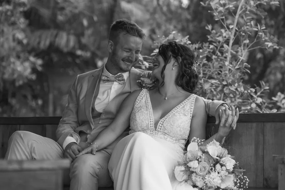 En noir et blanc, les mariés assis sur un banc rient ensemble, bouquet posé à côté