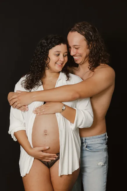Un couple enlacé en riant sur fond noir, la future maman en chemise blanche ouverte
