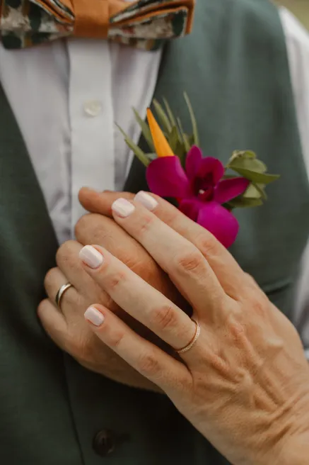 La main de la mariée, alliance au doigt, posée sur la boutonnière d’orchidée fuchsia du marié