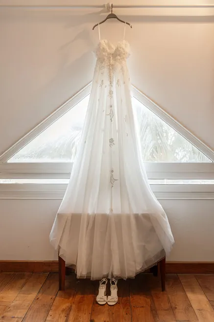 Robe de mariée en tulle suspendue devant une fenêtre triangulaire, escarpins posés dessous
