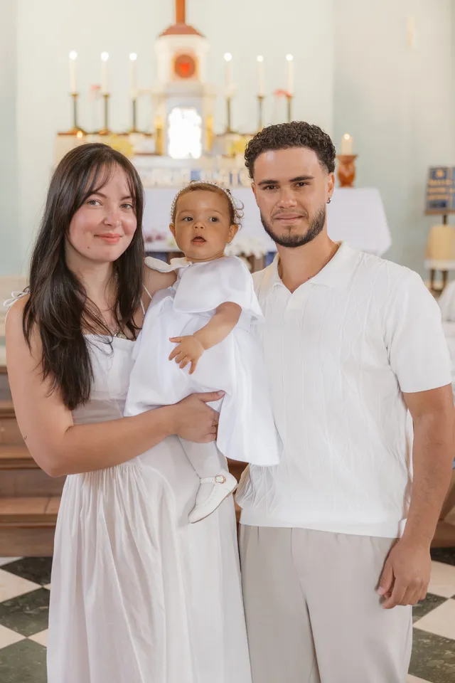 Un jeune couple en blanc tient son bébé à l’église, devant l’autel