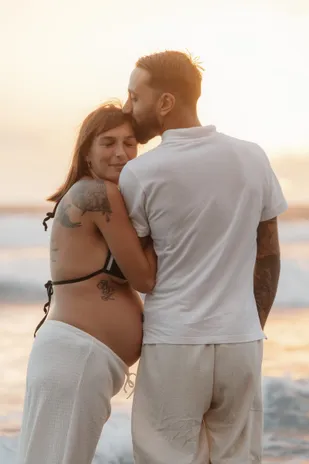Le futur papa embrasse le front de la future maman, dos à la mer au soleil couchant