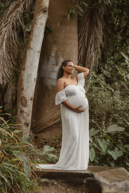 Future maman en robe blanche épaules nues, posée contre un tronc dans la végétation