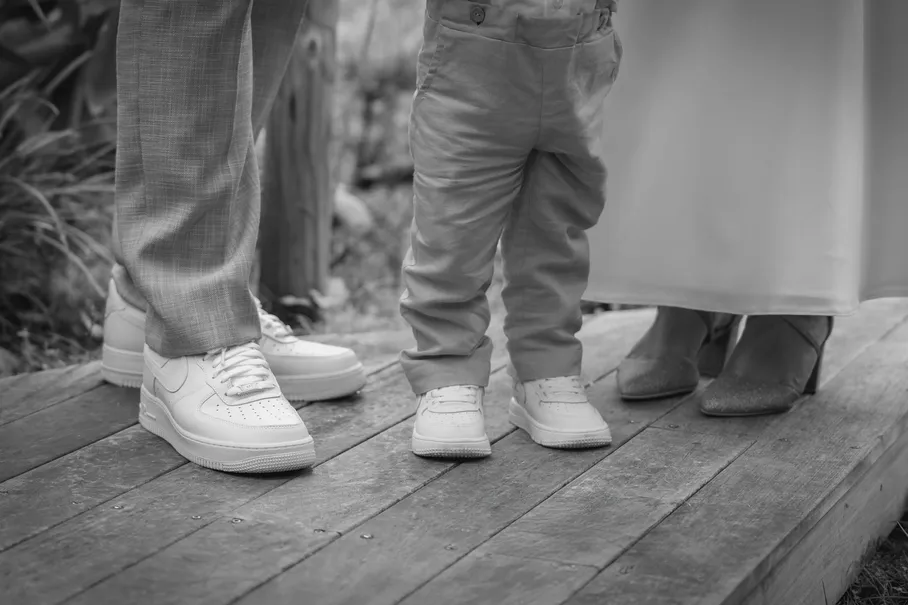 En noir et blanc, les pieds des mariés et d’un petit garçon en baskets blanches sur un ponton de bois