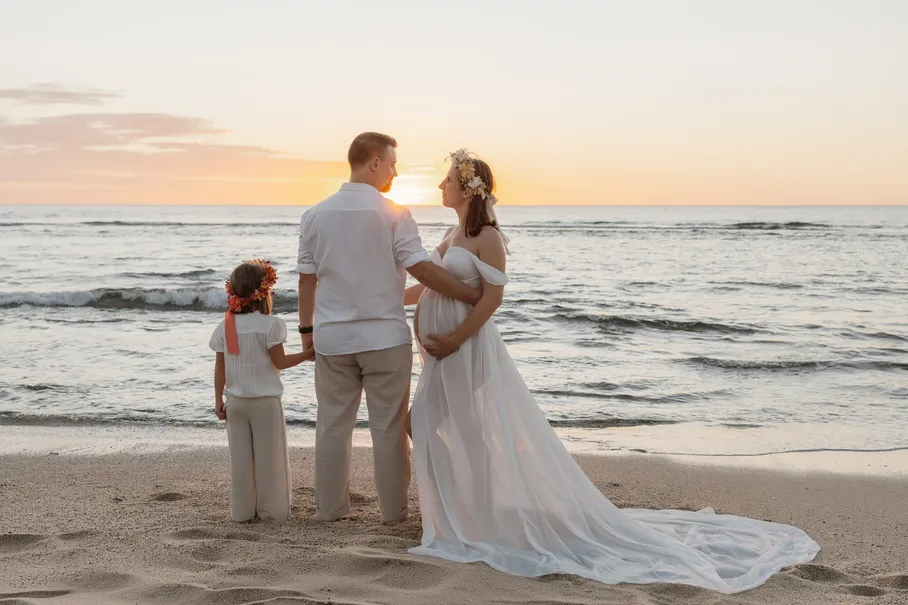 Une famille de dos regarde le soleil se coucher, la future maman en robe longue