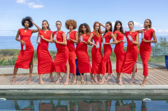 Dix mannequins en robes rouges alignées au bord d’une piscine, la mer derrière elles