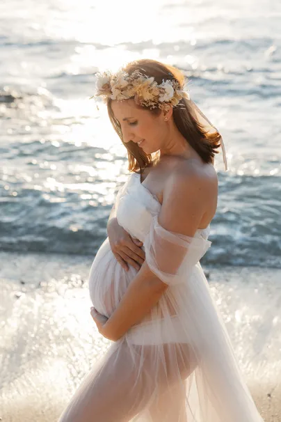 Future maman couronnée de fleurs séchées, tête baissée vers son ventre, mer scintillante derrière
