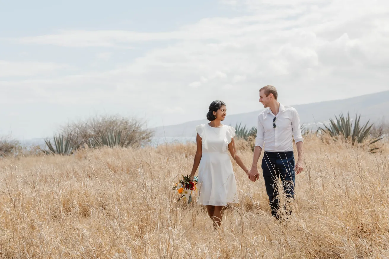 Un couple marche main dans la main dans les herbes sèches, elle en robe blanche avec son bouquet