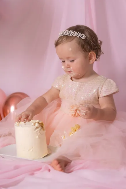 Une petite fille coiffée d’un diadème plonge les mains dans son gâteau blanc, assise dans le tulle rose