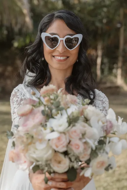 La mariée sourit avec des lunettes de soleil en cœur, bouquet de roses pâles en avant