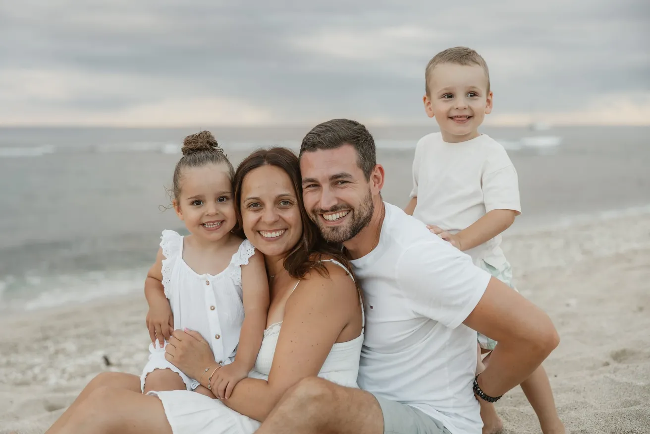 Une famille serrée sur le sable, les parents et leurs deux enfants souriants devant la mer