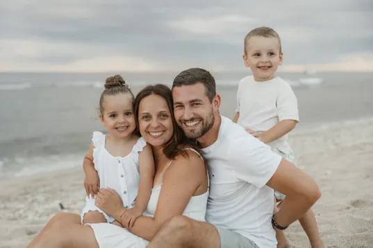 Une famille serrée sur le sable, les parents et leurs deux enfants souriants devant la mer