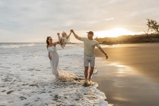 Des parents font tourner leur enfant à bout de bras, les pieds dans l’eau du couchant
