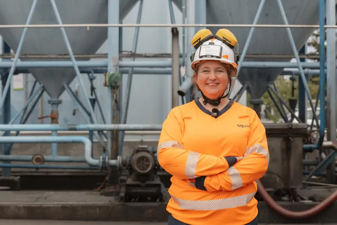 Une salariée en tenue orange et casque blanc sourit, bras croisés, devant les silos de son site