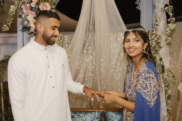 Le marié en tunique blanche passe la bague au doigt de la mariée en tenue bleue brodée