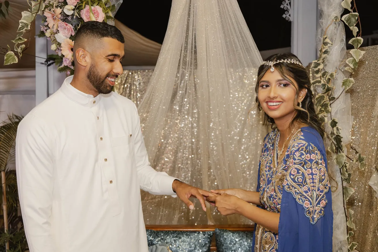 Le marié en tunique blanche passe la bague au doigt de la mariée en tenue bleue brodée