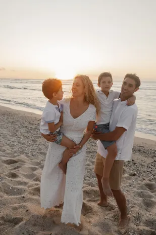 Une famille rit sur le sable face au couchant, chaque parent portant un petit garçon dans ses bras